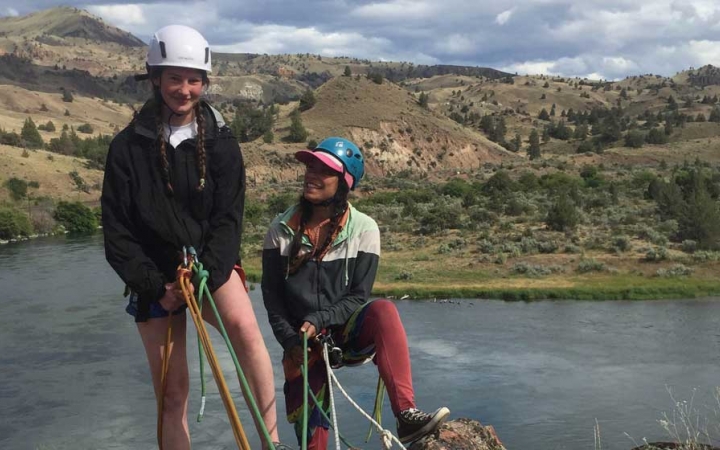 two people wearing rock climbing gear stand on a cliff about water before rappelling down